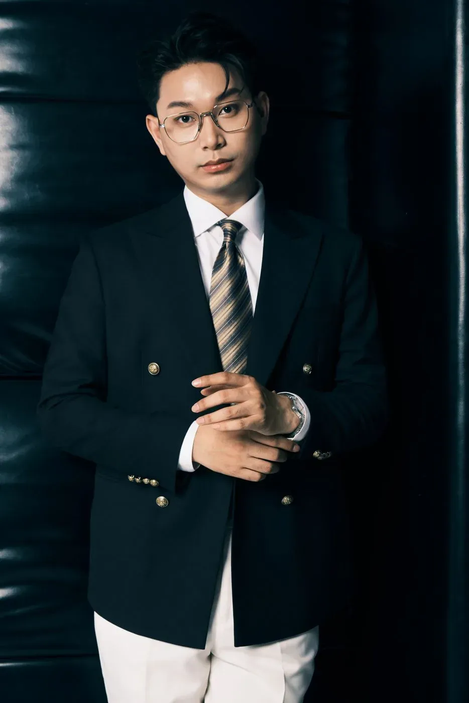 Chân dung MC Trọng Hiền mặc vest đen, đứng cạnh ghế da đen
